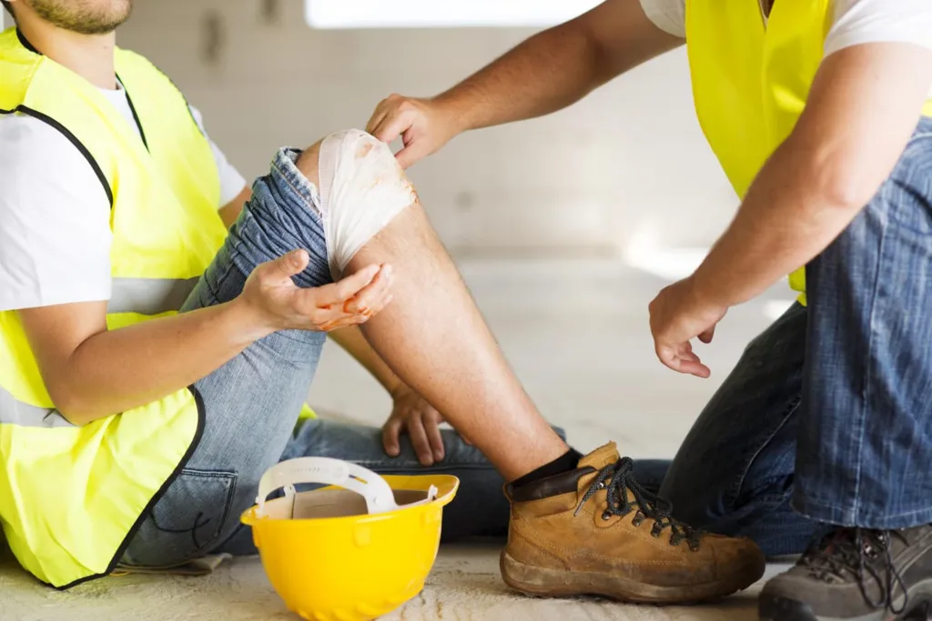 Indonesia Construction Safety Innovations: A worker in a safety vest receives first aid for a knee injury, with a bandage and some visible blood, while another person assists.