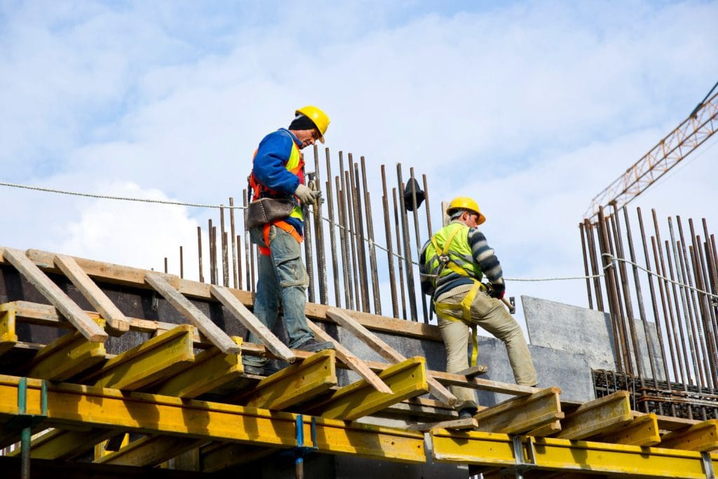 Indonesia Construction Safety Innovations: Two construction workers in safety gear and hard hats on scaffolding, working on a building structure with steel reinforcements.