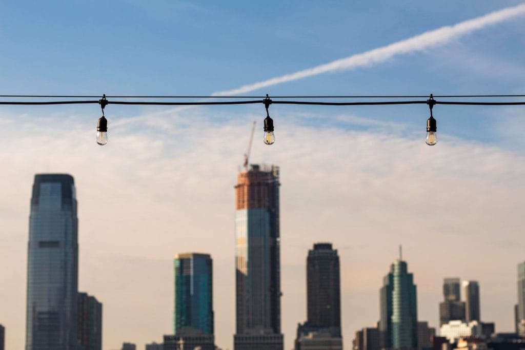 String lights hang on a wire against a blurred skyline featuring modern skyscrapers and a construction site under a blue sky to represent Energy-Efficient Building Designs.