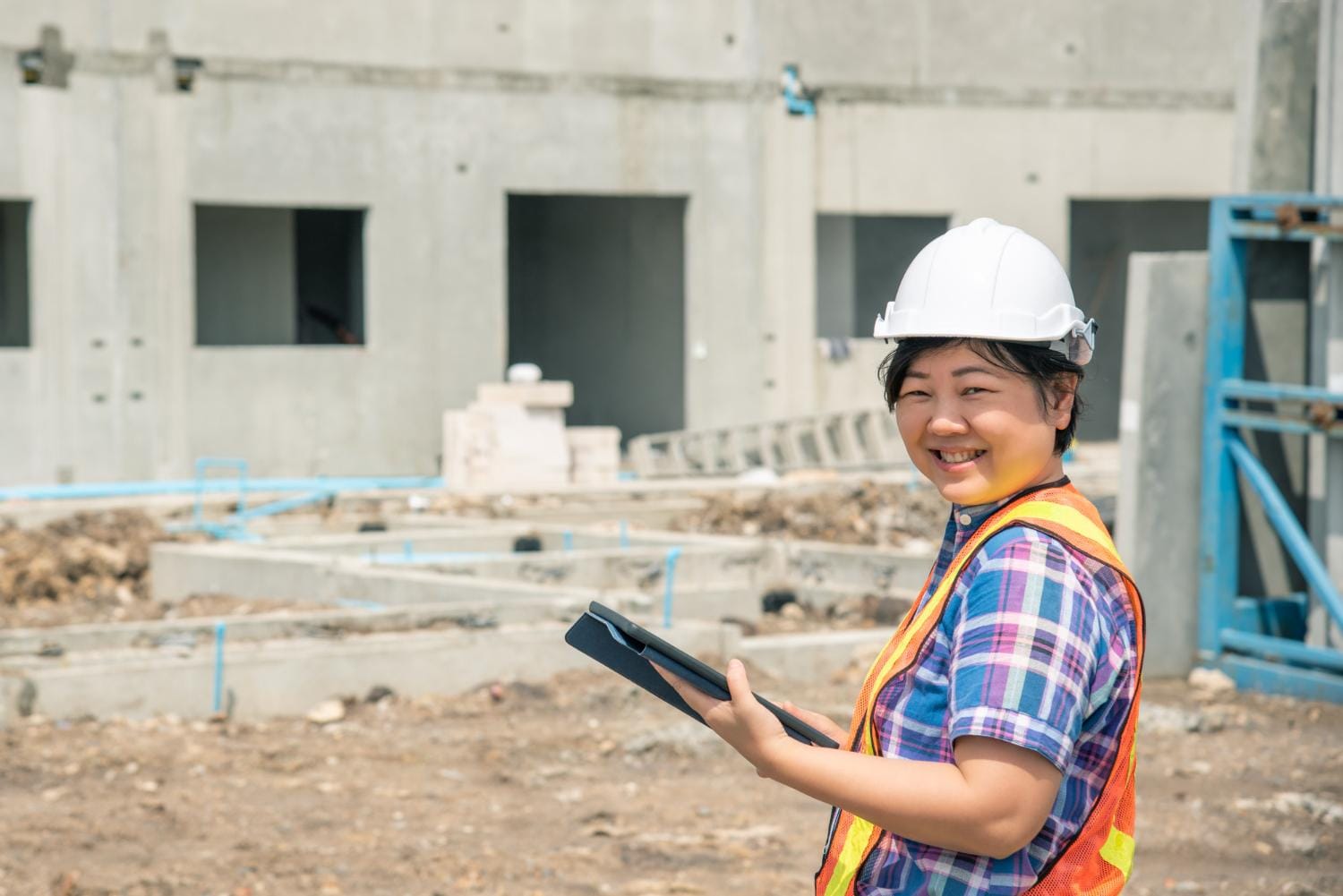 A construction worker in a hard hat and safety vest holds a tablet, overseeing a building site with concrete foundations and unfinished walls, representing Singapore Construction Cost Optimization.