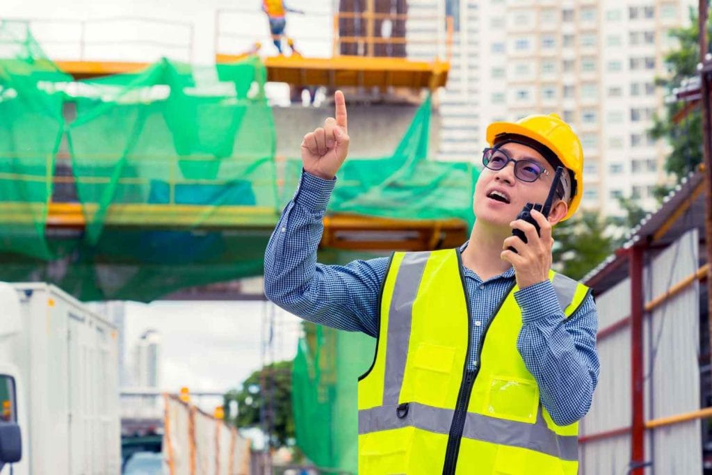 Singapore Digital Urban Planning Tools: A construction worker in a yellow safety vest and hard hat gestures while using a walkie-talkie at a construction site with scaffolding.