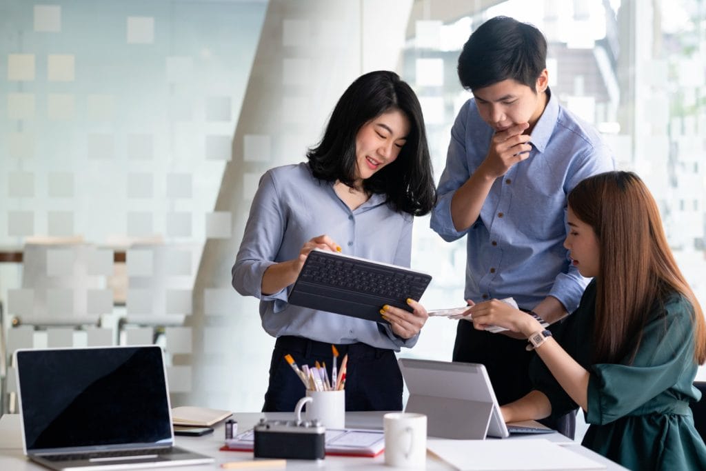 Eurogroup Consulting Southeast Asia: A diverse group of three professionals collaborating over tablets and laptops in a modern office setting, surrounded by stationary and paperwork.