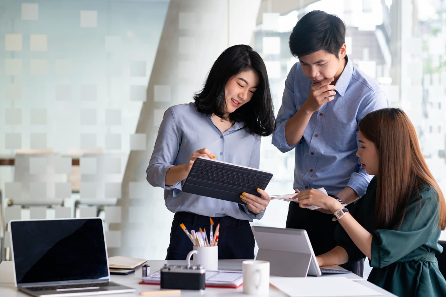 Eurogroup Consulting Southeast Asia: A diverse group of three professionals collaborating over tablets and laptops in a modern office setting, surrounded by stationary and paperwork.