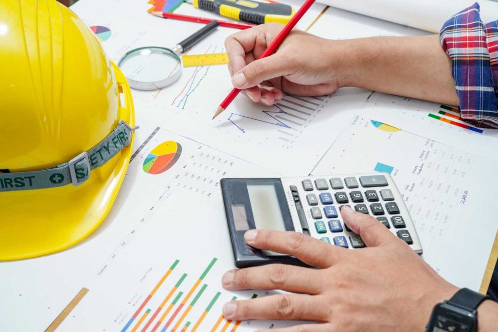 SEA Construction Cost Management: A person in a checked shirt calculates data on charts and graphs, with a yellow hard hat and tools nearby, emphasizing a construction context.