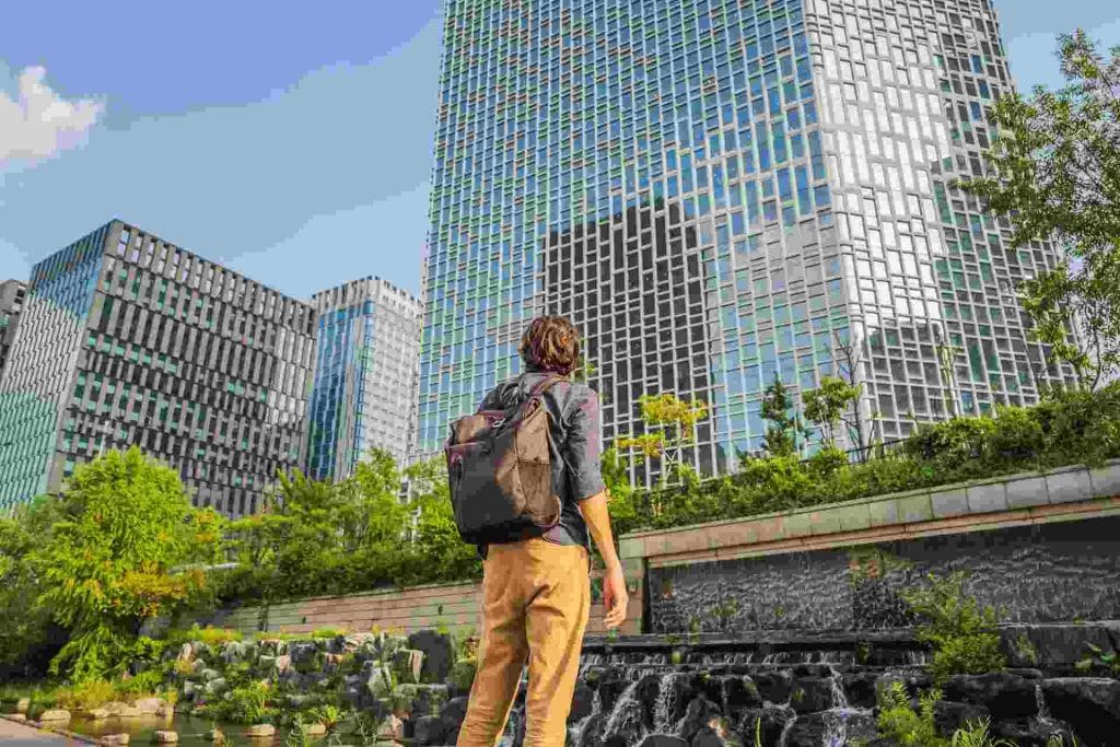 Philippines Urban Renewal Initiatives: A traveler gazes up at modern skyscrapers surrounded by greenery and a water feature in a vibrant city landscape.