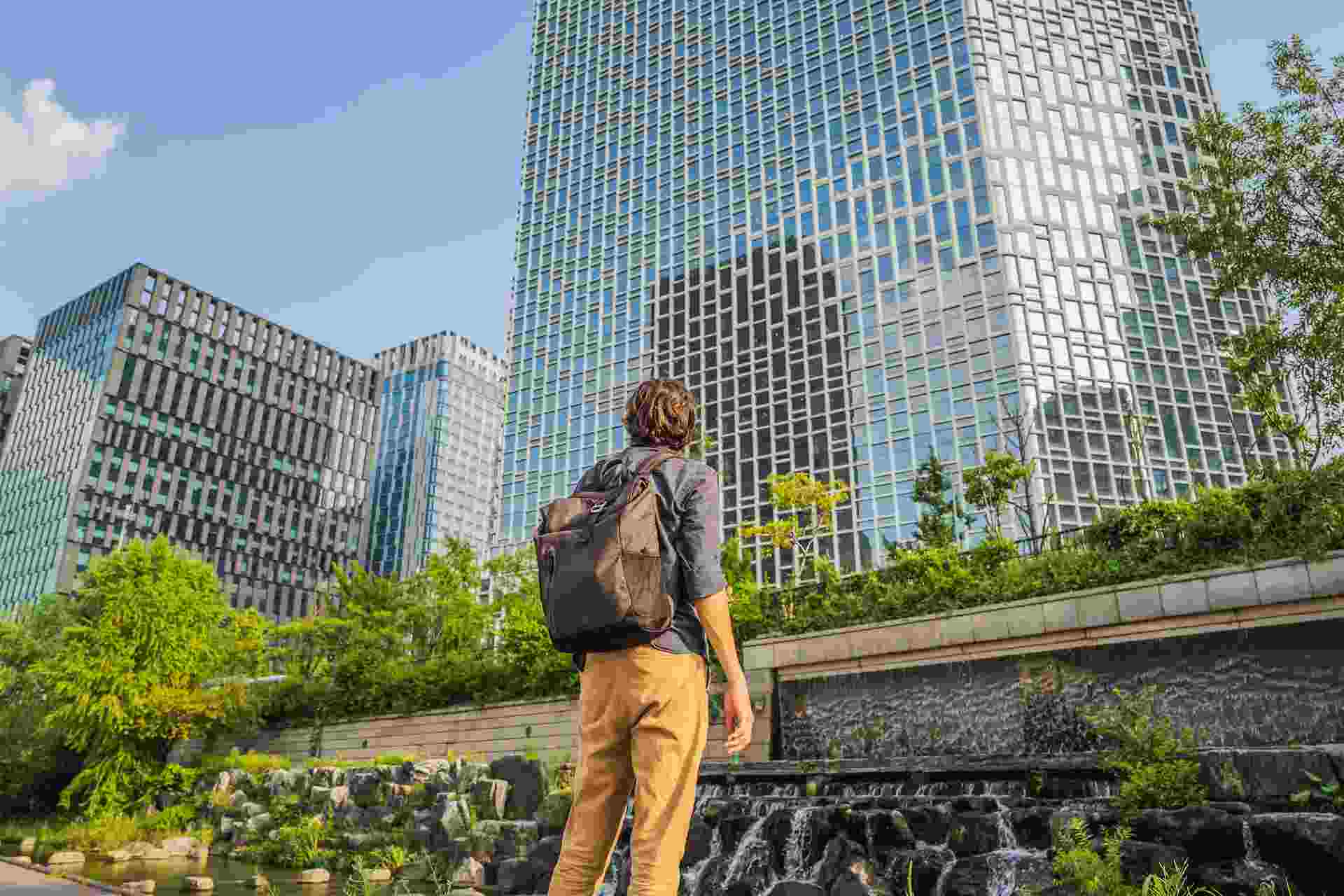 Philippines Urban Renewal Initiatives: A traveler gazes up at modern skyscrapers surrounded by greenery and a water feature in a vibrant city landscape.
