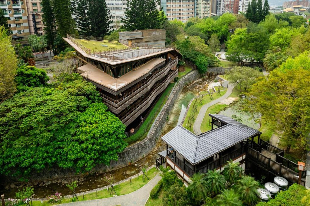 Philippines Energy-Efficient Building Designs: A modern wooden building with a green roof overlooks a lush park and winding pathways beside a small stream, framed by city buildings.