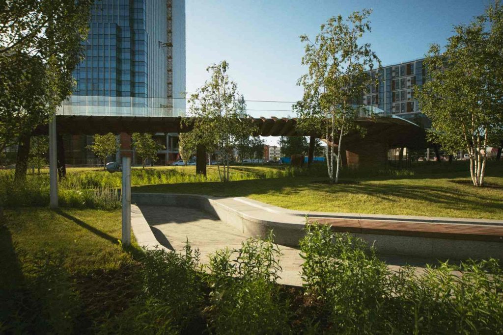 Malaysia Green Infrastructure Projects: A modern urban park featuring green grass, trees, and a unique walkway connecting two buildings under a clear blue sky.