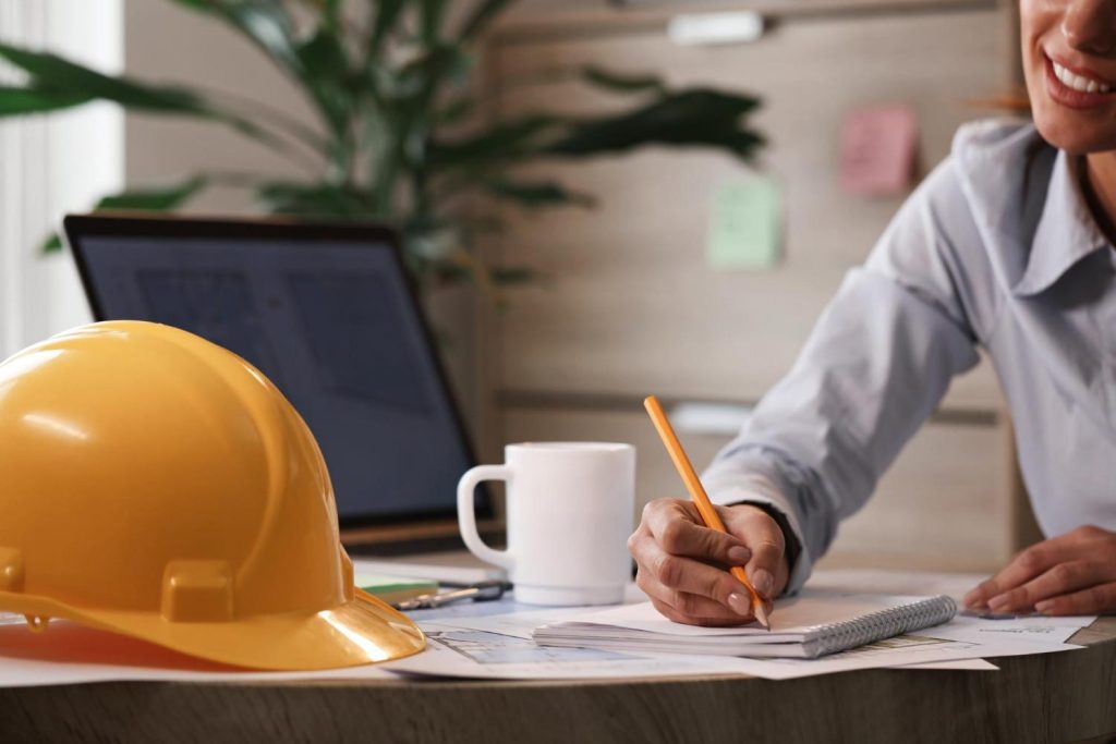 Philippines Construction Financing Challenges: A yellow hard hat sits beside a cup and scattered papers as a person writes notes, with a laptop in the background and plants nearby.