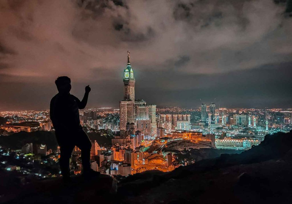 Saudi Vision 2030 progress: A silhouette of a person stands on a rocky outcrop, overlooking the illuminated city of Mecca and the clock tower at night.
