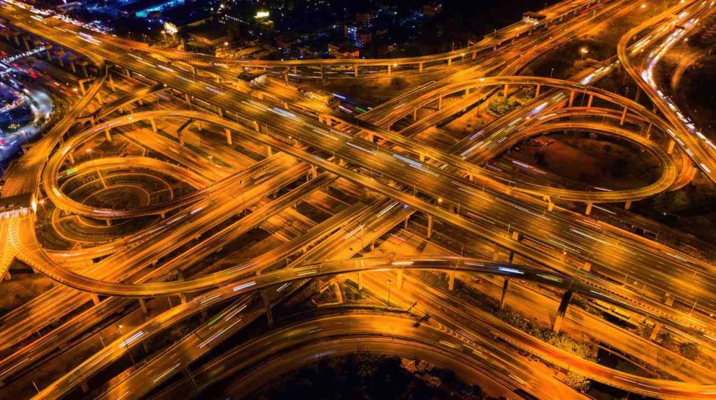 Malaysia Connectivity Infrastructure Projects: Aerial view of a bustling highway interchange at night, illuminated by orange street lights and moving traffic.