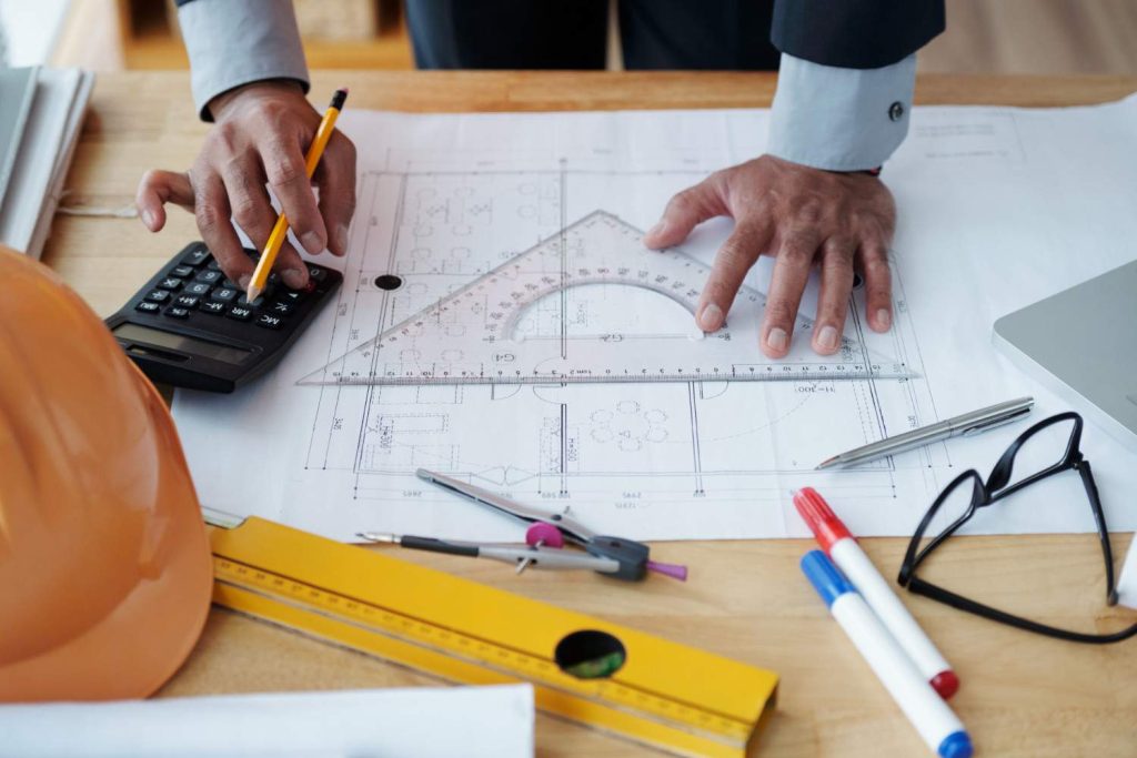 Vietnam Construction Cost Management: A person calculates measurements using a calculator and pencil on architectural blueprints, surrounded by drafting tools and a hard hat.