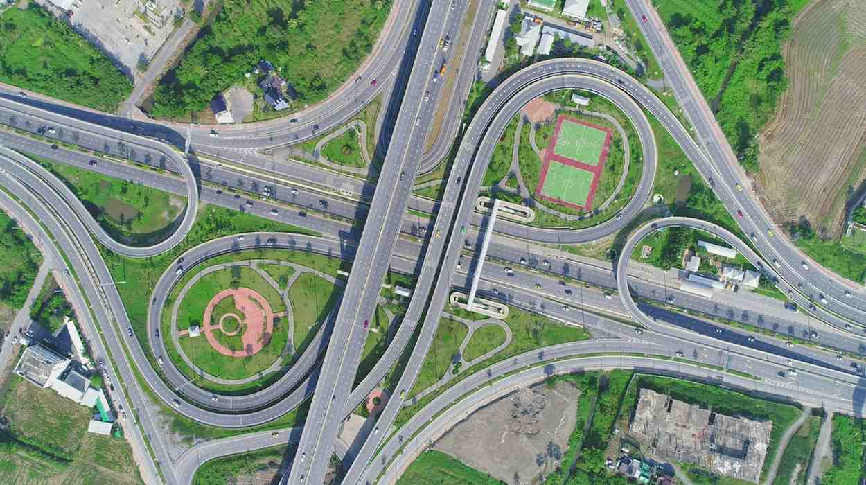 Indonesia Connectivity Infrastructure Projects: Aerial view of a complex highway interchange surrounded by greenery, with circular roads and adjacent sports fields.