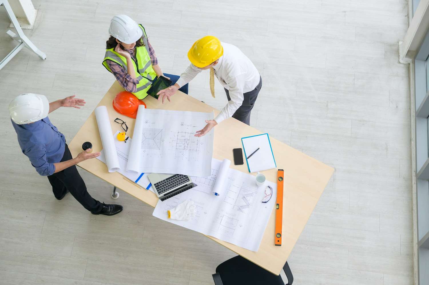 Indonesia Construction Cost Management: Two construction professionals in hard hats discuss blueprints at a table, with safety gear and a laptop visible.